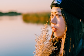 A person wearing a black beanie gazes thoughtfully across a calm body of water. The soft, golden light of the setting sun casts warm hues on their face and the fluffy plant they hold close. The background features blurred greenery and a serene, reflective water surface, creating a tranquil and contemplative scene.
