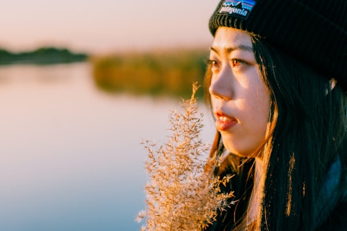 A person wearing a black beanie gazes thoughtfully across a calm body of water. The soft, golden light of the setting sun casts warm hues on their face and the fluffy plant they hold close. The background features blurred greenery and a serene, reflective water surface, creating a tranquil and contemplative scene.