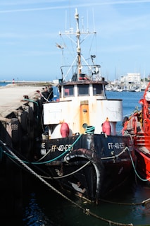A fishing boat moored at a harbor with ropes securing it to the dock. The vessel features antennas and communication equipment on top, and the hull displays signs of rust. Several other boats are visible in the background along with a clear blue sky.