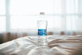 A clear plastic water bottle with a blue label, marked with the Aquafina brand, rests on a rumpled white fabric surface, possibly a bed. The background is bright and softly blurred, with natural light streaming through a window, creating a serene and minimalistic atmosphere.