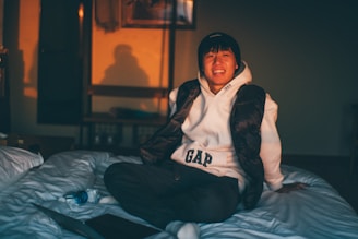 A person wearing a white hoodie and black vest is sitting on a bed, smiling warmly. The room is softly lit by warm, natural light, casting shadows on the wall. Nearby, a blue water bottle and a closed laptop are placed on the bed.
