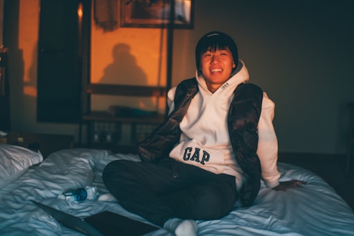 A person wearing a white hoodie and black vest is sitting on a bed, smiling warmly. The room is softly lit by warm, natural light, casting shadows on the wall. Nearby, a blue water bottle and a closed laptop are placed on the bed.