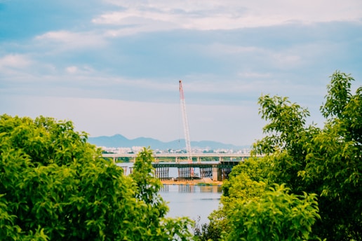A picturesque view of a bridge under construction over a river, surrounded by lush green trees. The crane in the middle is prominently standing against a backdrop of distant mountains and a cloudy sky.