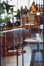 A cozy indoor setting featuring a neatly arranged dining table with stacked plates, utensils, and condiments. Warm, ambient lighting is provided by several copper pendant lamps. Indoor plants contribute to the decor, alongside a leather chair and string lights hanging in the foreground.