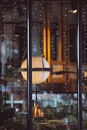 A large spherical lamp suspended within a glass enclosure, surrounded by strings of delicate fairy lights. It appears to be part of a cozy interior setting with warm wooden tones and reflections on the glass. Behind the lamp, shelves with various items and some plants can be seen, suggesting an inviting atmosphere.