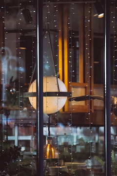 A large spherical lamp suspended within a glass enclosure, surrounded by strings of delicate fairy lights. It appears to be part of a cozy interior setting with warm wooden tones and reflections on the glass. Behind the lamp, shelves with various items and some plants can be seen, suggesting an inviting atmosphere.