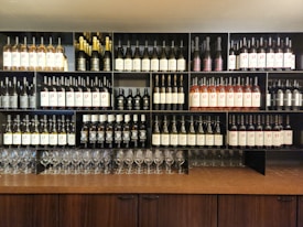 A wooden cabinet displays various bottles of wine organized neatly on shelves. The shelves hold different kinds of wine, including white, red, and sparkling varieties. Wine glasses are arranged in rows at the bottom, adding to the elegant arrangement.