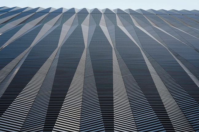 A geometric pattern covers the surface of a building. The structure displays a series of repeating triangular shapes, creating a dynamic zigzag effect. The design is uniform and symmetrical, with lines converging at the top, enhancing the structure's height and depth. The surface reflects light subtly, emphasizing the contrast between the light and shadowed areas.
