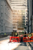 A city street scene with a garbage truck in the center of the image. The truck is surrounded by workers wearing high-visibility vests and helmets. They are seen handling waste and operating around the truck, which is behind red and orange barriers. The street is flanked by tall buildings, with modern glass structures visible in the background. The lighting suggests early morning or late afternoon, casting a warm, soft glow over the scene.