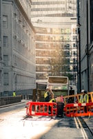 A city street scene with a garbage truck in the center of the image. The truck is surrounded by workers wearing high-visibility vests and helmets. They are seen handling waste and operating around the truck, which is behind red and orange barriers. The street is flanked by tall buildings, with modern glass structures visible in the background. The lighting suggests early morning or late afternoon, casting a warm, soft glow over the scene.