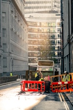 A city street scene with a garbage truck in the center of the image. The truck is surrounded by workers wearing high-visibility vests and helmets. They are seen handling waste and operating around the truck, which is behind red and orange barriers. The street is flanked by tall buildings, with modern glass structures visible in the background. The lighting suggests early morning or late afternoon, casting a warm, soft glow over the scene.