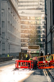 A city street scene with a garbage truck in the center of the image. The truck is surrounded by workers wearing high-visibility vests and helmets. They are seen handling waste and operating around the truck, which is behind red and orange barriers. The street is flanked by tall buildings, with modern glass structures visible in the background. The lighting suggests early morning or late afternoon, casting a warm, soft glow over the scene.