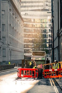 A city street scene with a garbage truck in the center of the image. The truck is surrounded by workers wearing high-visibility vests and helmets. They are seen handling waste and operating around the truck, which is behind red and orange barriers. The street is flanked by tall buildings, with modern glass structures visible in the background. The lighting suggests early morning or late afternoon, casting a warm, soft glow over the scene.