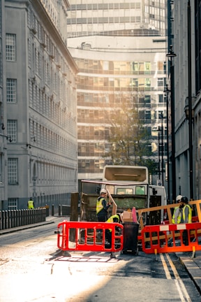 A city street scene with a garbage truck in the center of the image. The truck is surrounded by workers wearing high-visibility vests and helmets. They are seen handling waste and operating around the truck, which is behind red and orange barriers. The street is flanked by tall buildings, with modern glass structures visible in the background. The lighting suggests early morning or late afternoon, casting a warm, soft glow over the scene.