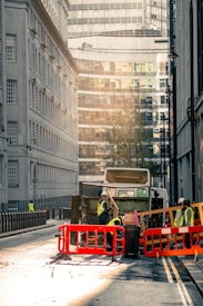 A city street scene with a garbage truck in the center of the image. The truck is surrounded by workers wearing high-visibility vests and helmets. They are seen handling waste and operating around the truck, which is behind red and orange barriers. The street is flanked by tall buildings, with modern glass structures visible in the background. The lighting suggests early morning or late afternoon, casting a warm, soft glow over the scene.