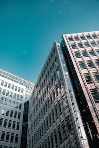 Tall, modern office buildings with reflective glass and structured facades reach up towards a clear blue sky. The architectural design showcases geometric patterns and symmetry.