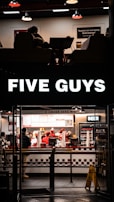 A fast-food restaurant scene with several people wearing uniforms behind the counter, serving customers. The counter displays various menu options, and a bright sign with the brand name is illuminated above the entrance. The interior features a clean, modern design with red and white accents, and a seating area partially visible.