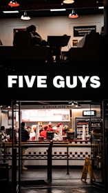A fast-food restaurant scene with several people wearing uniforms behind the counter, serving customers. The counter displays various menu options, and a bright sign with the brand name is illuminated above the entrance. The interior features a clean, modern design with red and white accents, and a seating area partially visible.