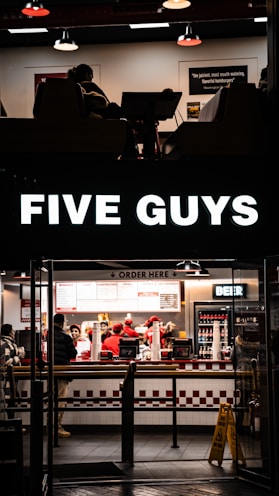 A fast-food restaurant scene with several people wearing uniforms behind the counter, serving customers. The counter displays various menu options, and a bright sign with the brand name is illuminated above the entrance. The interior features a clean, modern design with red and white accents, and a seating area partially visible.