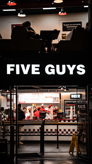 A fast-food restaurant scene with several people wearing uniforms behind the counter, serving customers. The counter displays various menu options, and a bright sign with the brand name is illuminated above the entrance. The interior features a clean, modern design with red and white accents, and a seating area partially visible.