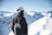 A person wearing a black jacket and white helmet stands overlooking a snowy mountain range. The helmet has 'APRES' written on it, and the jacket features a logo with 'THE SNOW EXPERIENCE DAVOS'. The scene is bright with snow-capped peaks under a clear blue sky.