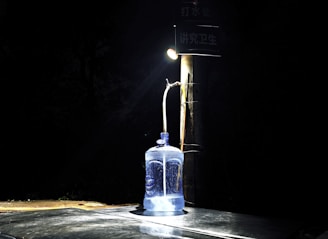 A large blue water container is placed on a dark surface, illuminated by a single overhead light attached to a pole. The background is very dark, creating a stark contrast with the lit container. The setup suggests an outdoor environment with sparse lighting.