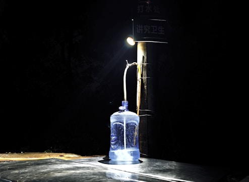A large blue water container is placed on a dark surface, illuminated by a single overhead light attached to a pole. The background is very dark, creating a stark contrast with the lit container. The setup suggests an outdoor environment with sparse lighting.