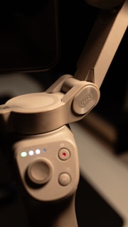 A close-up view of a gray electronic device featuring a hinge mechanism and buttons. The device has a smooth texture and displays multiple colored lights indicating its status. The word 'OSMO' is embossed on one part of the hinge.