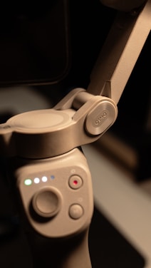 A close-up view of a gray electronic device featuring a hinge mechanism and buttons. The device has a smooth texture and displays multiple colored lights indicating its status. The word 'OSMO' is embossed on one part of the hinge.