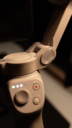 A close-up view of a gray electronic device featuring a hinge mechanism and buttons. The device has a smooth texture and displays multiple colored lights indicating its status. The word 'OSMO' is embossed on one part of the hinge.