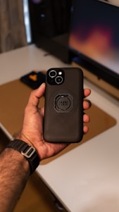 A hand holds a smartphone with a black case that has a circular mount in the center. The background includes a blurred computer monitor with warm colors and a desk with various items.