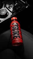 A bottle of red hydration drink with the label 'Tropical Punch' and 'PRIME' printed in large black letters is resting on a dark car seat. The bottle is partially illuminated, drawing attention to its vibrant color amidst the dimly lit interior.