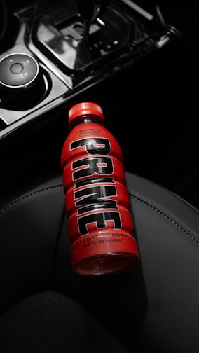 A bottle of red hydration drink with the label 'Tropical Punch' and 'PRIME' printed in large black letters is resting on a dark car seat. The bottle is partially illuminated, drawing attention to its vibrant color amidst the dimly lit interior.