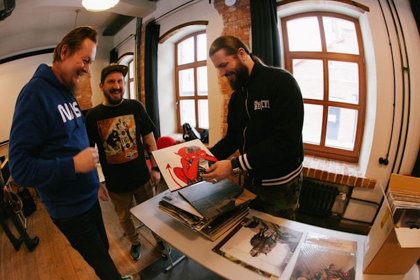 Three men are gathered around a table with stacks of vinyl records. One man is holding an album with a red devil illustration on the cover while the other two look on, smiling. The room has brick walls and large windows letting in natural light.