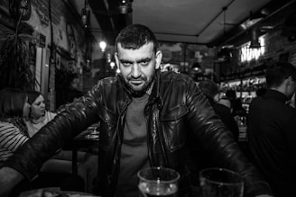 A man with a beard and a serious expression stands in a bustling bar, wearing a leather jacket. Seated patrons are visible in the background, with dim lighting creating a cozy atmosphere.