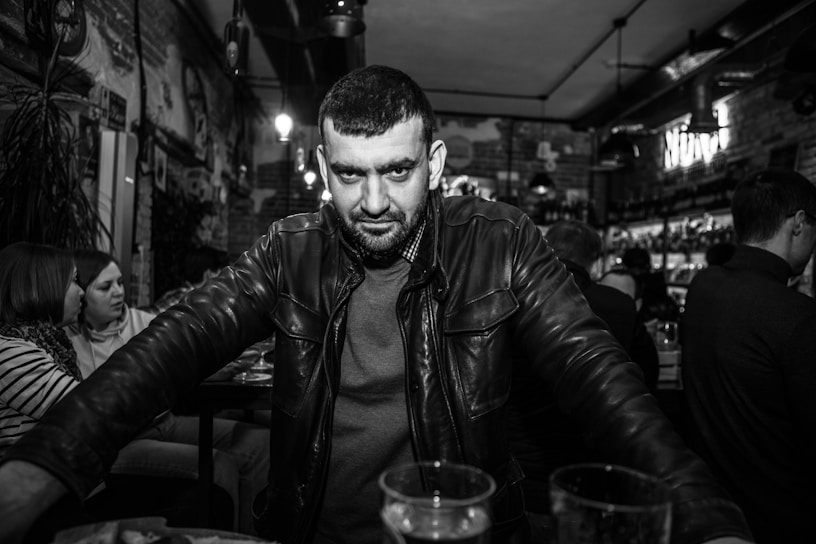 A man with a beard and a serious expression stands in a bustling bar, wearing a leather jacket. Seated patrons are visible in the background, with dim lighting creating a cozy atmosphere.