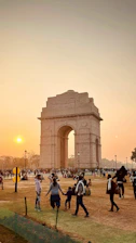 The majestic India Gate glowing warmly at sunset with visitors strolling nearby.