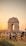 Appearing in the image is a large monumental arch known as India Gate, situated in an open area with a number of people walking and gathering around it. The sun is setting, casting an orange glow across the sky and landscape. The ground is mostly grassy with some bare patches, and there are street lamps and barriers visible.