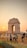 Appearing in the image is a large monumental arch known as India Gate, situated in an open area with a number of people walking and gathering around it. The sun is setting, casting an orange glow across the sky and landscape. The ground is mostly grassy with some bare patches, and there are street lamps and barriers visible.