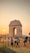 Appearing in the image is a large monumental arch known as India Gate, situated in an open area with a number of people walking and gathering around it. The sun is setting, casting an orange glow across the sky and landscape. The ground is mostly grassy with some bare patches, and there are street lamps and barriers visible.