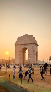 Appearing in the image is a large monumental arch known as India Gate, situated in an open area with a number of people walking and gathering around it. The sun is setting, casting an orange glow across the sky and landscape. The ground is mostly grassy with some bare patches, and there are street lamps and barriers visible.