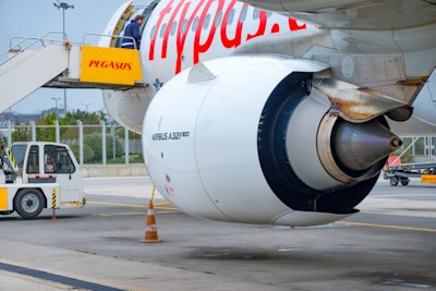 A close-up view of an airplane engine on the tarmac, with a visible Airbus A321neo label and a boarding stairway with passengers. A ground service vehicle is also present, along with a traffic cone on the ground. The scene occurs at an airport with a fence and some greenery in the background.