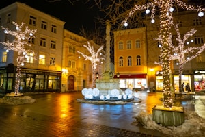 A beautifully decorated town square at night with trees adorned in glowing string lights and illuminated orbs. The scene showcases a central fountain surrounded by charming old buildings. A few patches of snow are visible on the ground, and the street reflects warm lights from nearby shops and cafes.