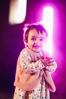 A young child is smiling while clapping hands in an indoor setting with vibrant lighting. The child is wearing a colorful patterned outfit with a pink fur vest, and there is a bright pinkish-purple light behind creating a glowing effect.