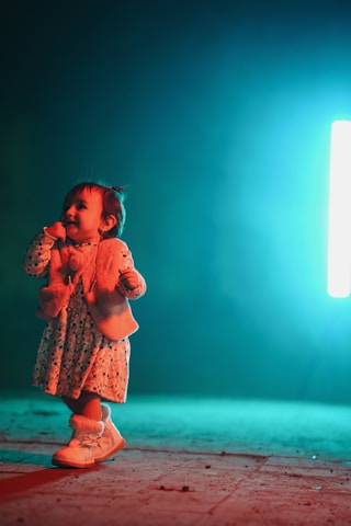 A young child wearing a floral dress, a fluffy vest, and boots stands cheerfully against a dramatic teal and dark background. The lighting creates a vibrant contrast, highlighting the joyful expression of the child.