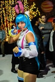 A person dressed in vibrant cosplay with blue hair, pink and blue accessories, and a playful pose. The background is filled with warm, glowing lights and other individuals in casual and costume attire.