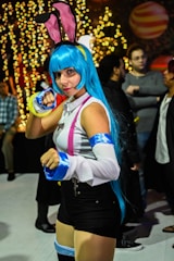 A person dressed in vibrant cosplay with blue hair, pink and blue accessories, and a playful pose. The background is filled with warm, glowing lights and other individuals in casual and costume attire.