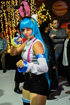 A person dressed in vibrant cosplay with blue hair, pink and blue accessories, and a playful pose. The background is filled with warm, glowing lights and other individuals in casual and costume attire.