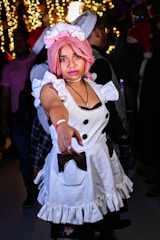 A person with pink hair is dressed in a maid costume, featuring a white dress with black buttons and ruffled edges. The background is festive, with blurred decorative lights and other people visible, some wearing Santa hats.