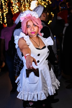 A person with pink hair is dressed in a maid costume, featuring a white dress with black buttons and ruffled edges. The background is festive, with blurred decorative lights and other people visible, some wearing Santa hats.